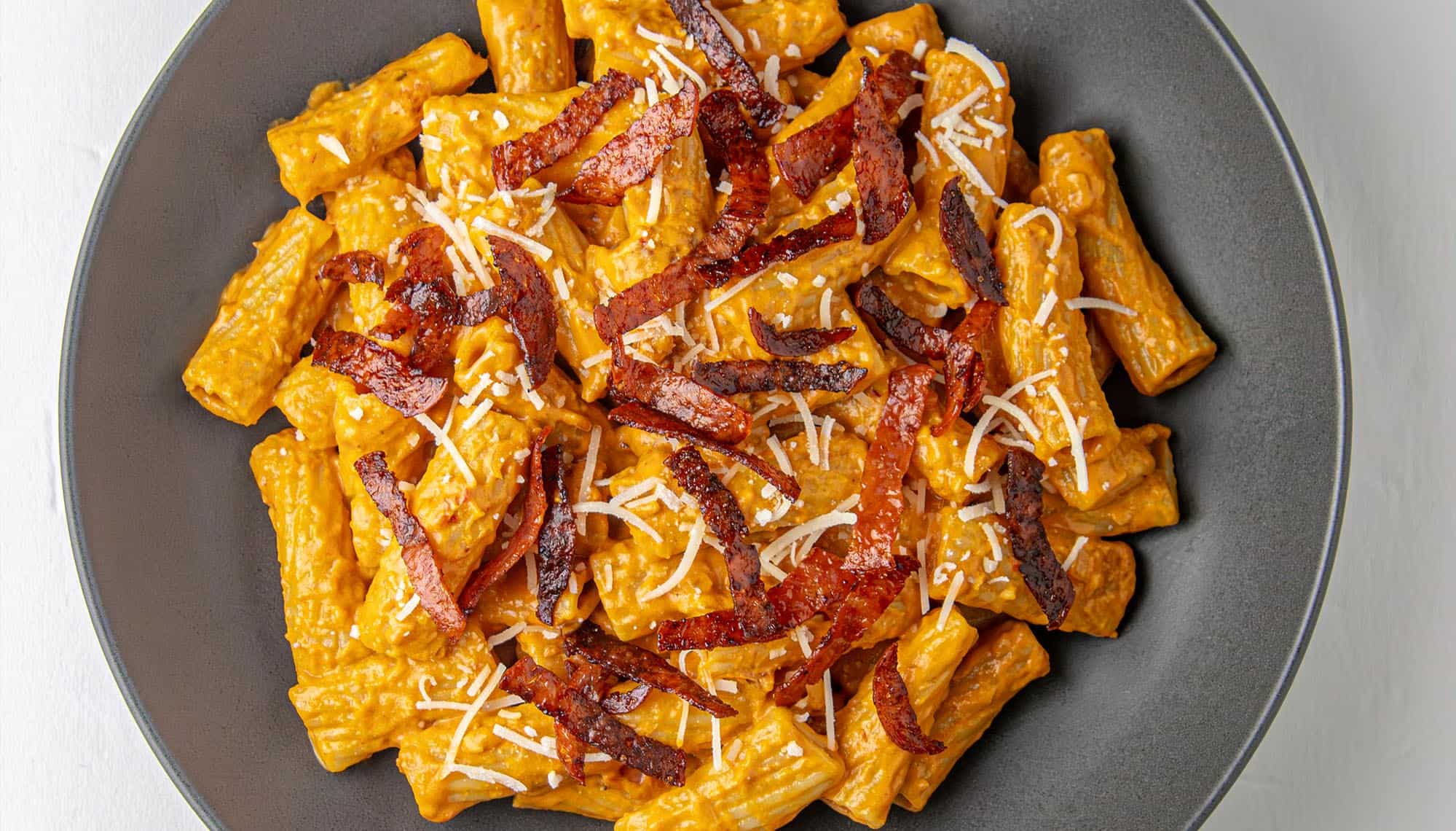 Rigatoni pasta in creamy orange sauce topped with crispy bacon strips and grated cheese in a dark bowl.