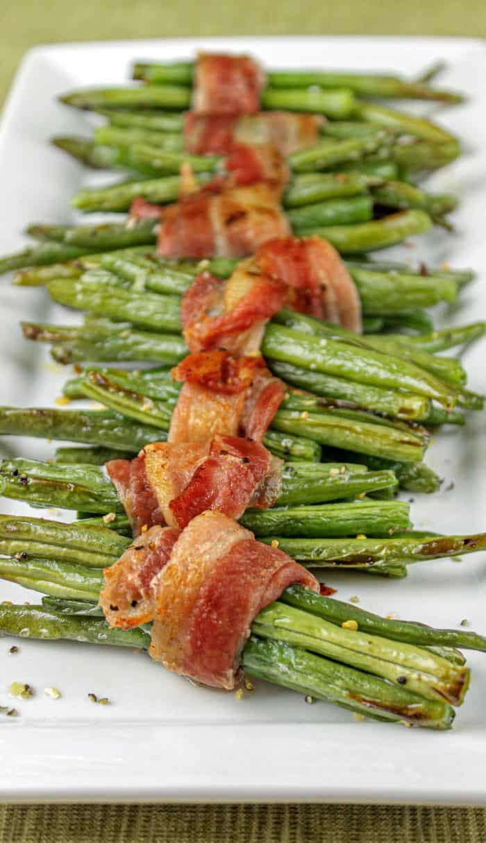 Green bean bundles wrapped in crispy cooked bacon on a white rectangular plate.