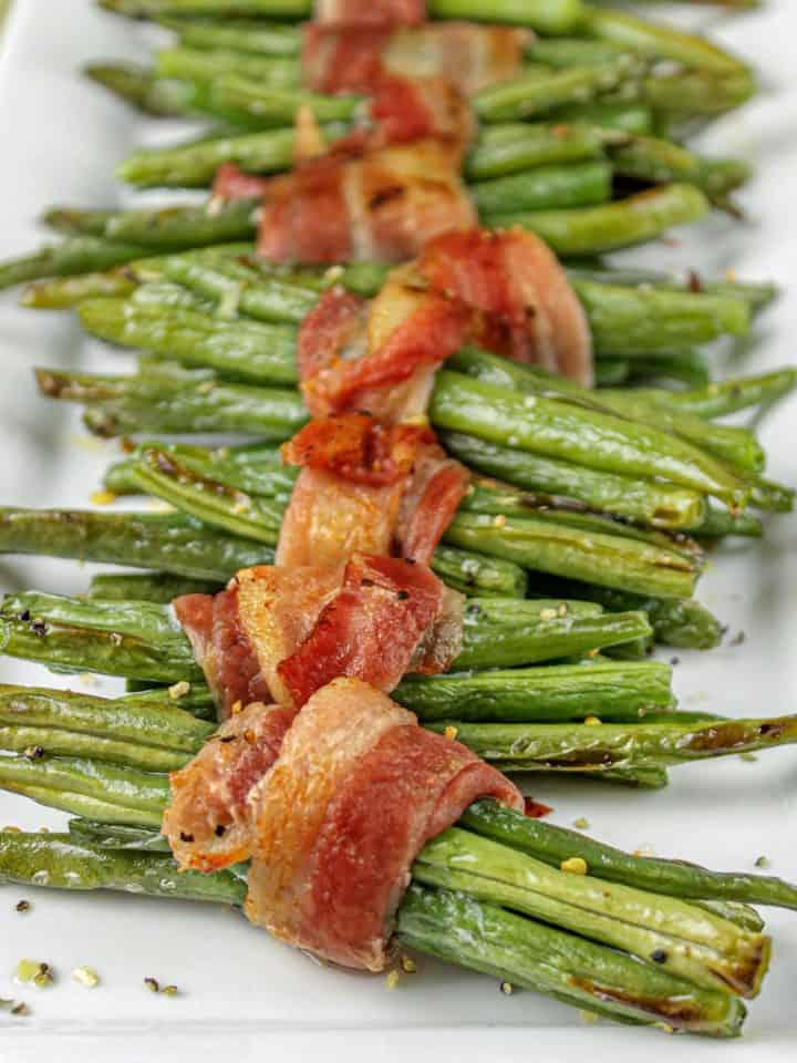 Green bean bundles wrapped in crispy cooked bacon on a white rectangular plate.