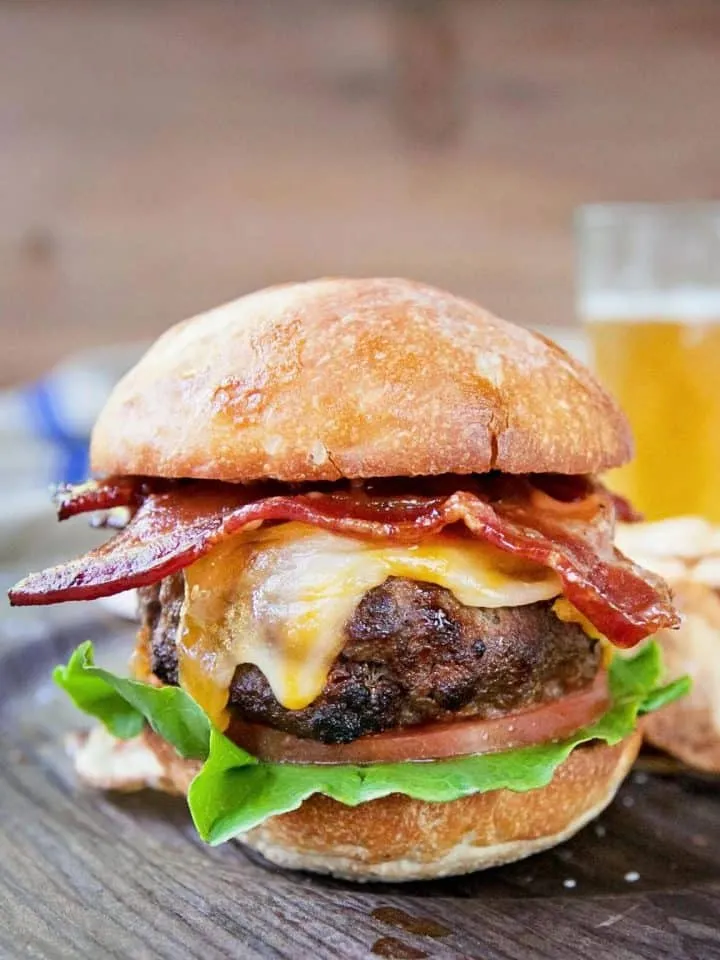 Bacon cheeseburger with lettuce and tomato on a toasted bun, served on a wooden plate with a glass of beer in the background.