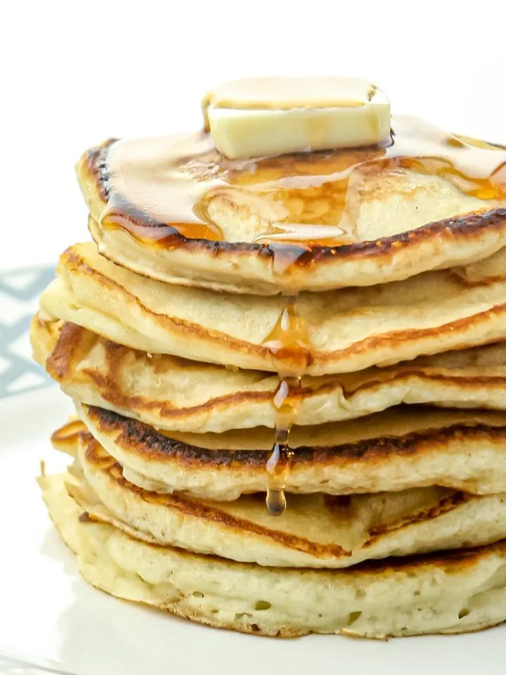 Stack of thick golden pancakes with melting butter and syrup dripping down on a white plate.