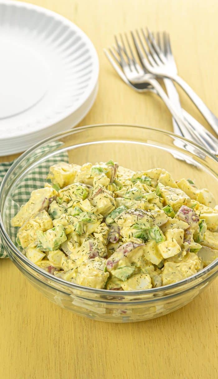 Bowl of creamy yellow potato salad with celery, red-skinned potatoes, dill, and green onions on a yellow wooden table.