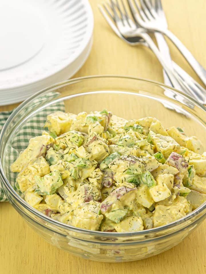 Bowl of creamy yellow potato salad with celery, red-skinned potatoes, dill, and green onions on a yellow wooden table.
