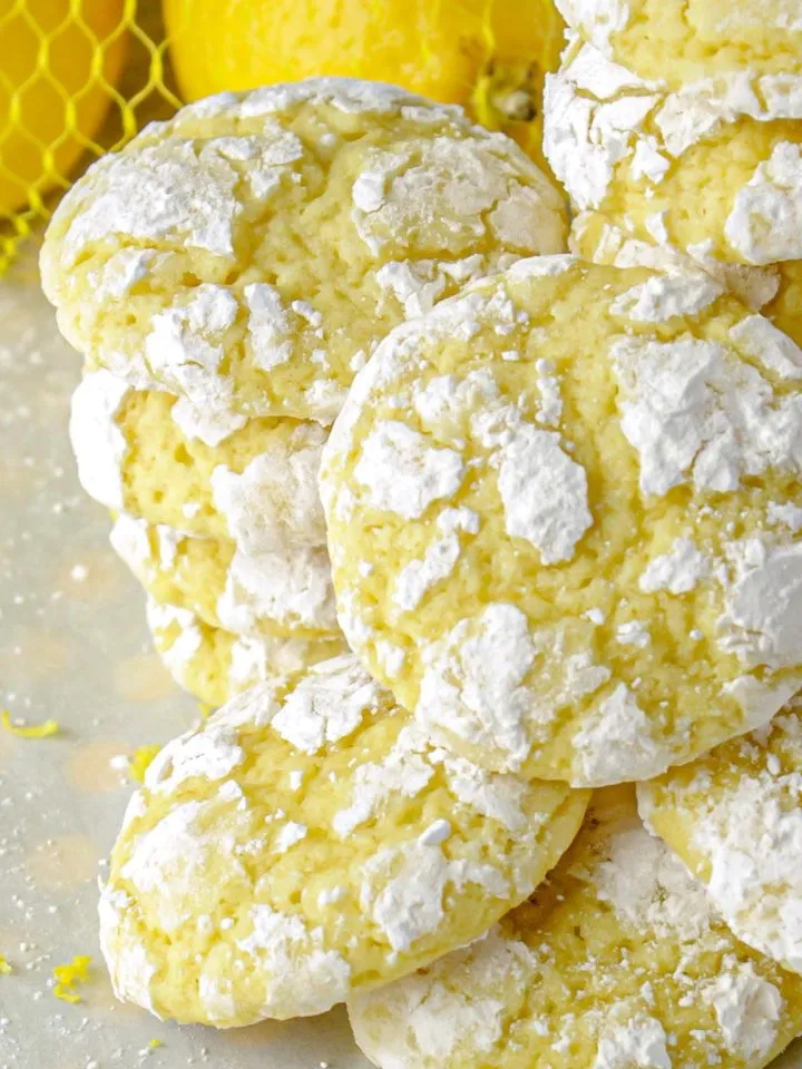 Crinkled lemon cookies dusted with powdered sugar stacked on a surface with whole lemons in the background.