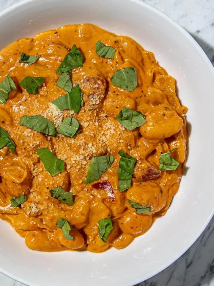 Gnocchi in creamy palomino sauce topped with chopped basil and grated Parmesan in a white bowl on marble surface.