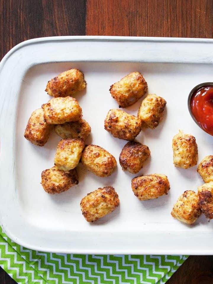 Golden-brown crispy tater tots on a white tray with a side of red ketchup, placed on a green and white zigzag patterned napkin.