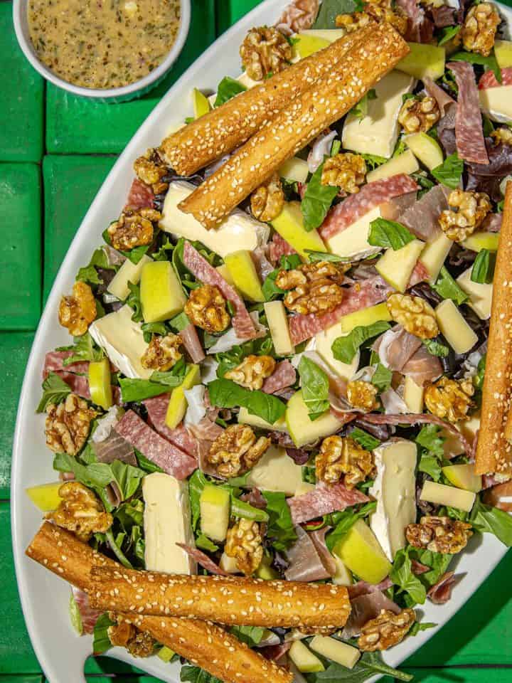Salad with mixed greens, brie cheese, salami, walnuts, apple slices, and breadsticks topped with sesame seeds, served with a herb dressing.