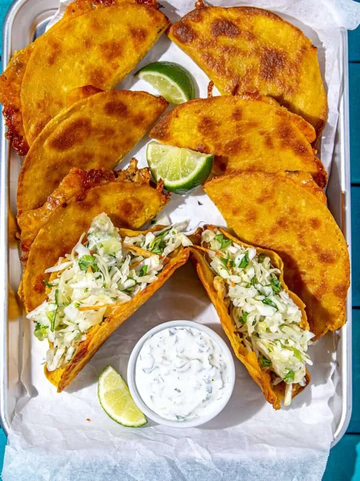 Crispy fried tacos and two soft tacos with shredded cabbage, lime wedges, and a bowl of creamy sauce on a metal tray lined with parchment paper.