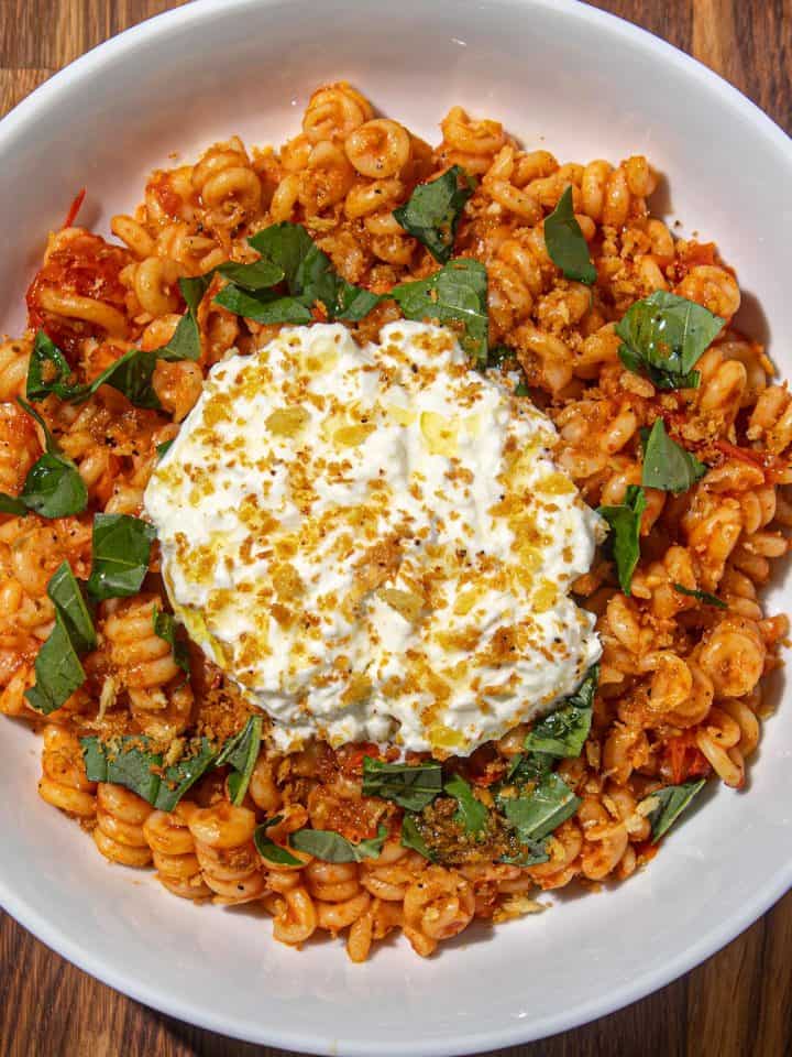 Fusilli corti pasta with tomato sauce, fresh basil, Burrata cheese, and breadcrumb topping in a white bowl on a wooden surface.