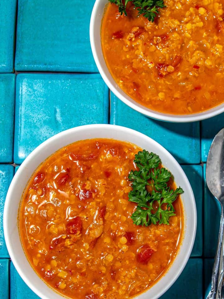Two bowls of red lentil soup garnished with parsley on blue tiled surface with a spoon beside them.