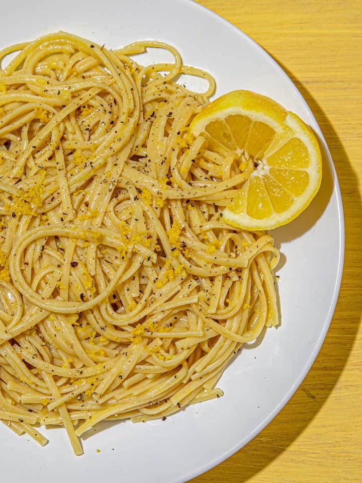 Linguine pasta seasoned with black pepper and lemon zest, garnished with a lemon wedge on a white plate on a yellow wooden surface.