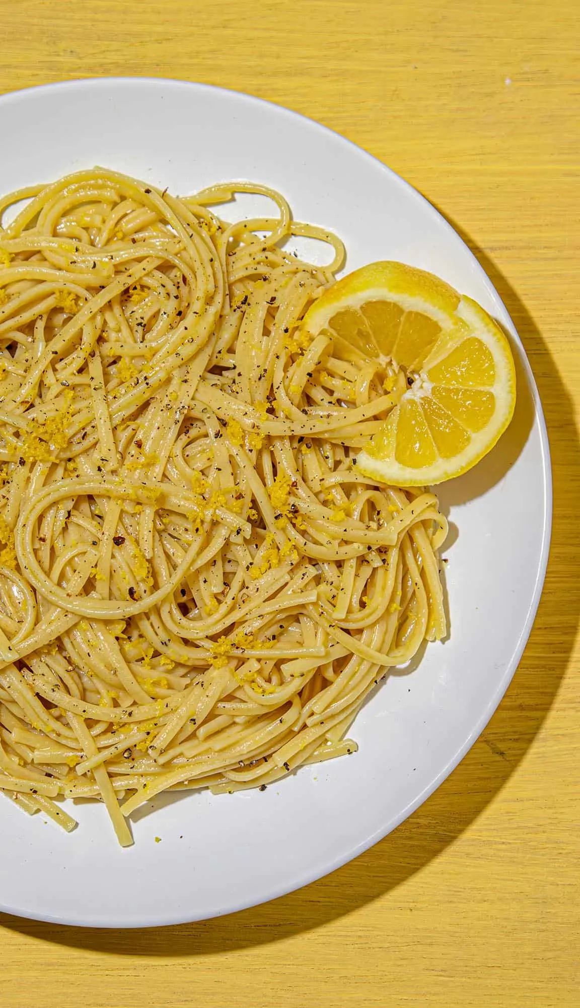 Linguine pasta seasoned with black pepper and lemon zest, garnished with a lemon wedge on a white plate on a yellow wooden surface.