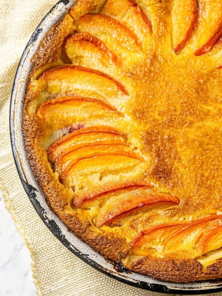 Golden brown baked tart with peach slices in a round black rimmed dish on a cream textured cloth.