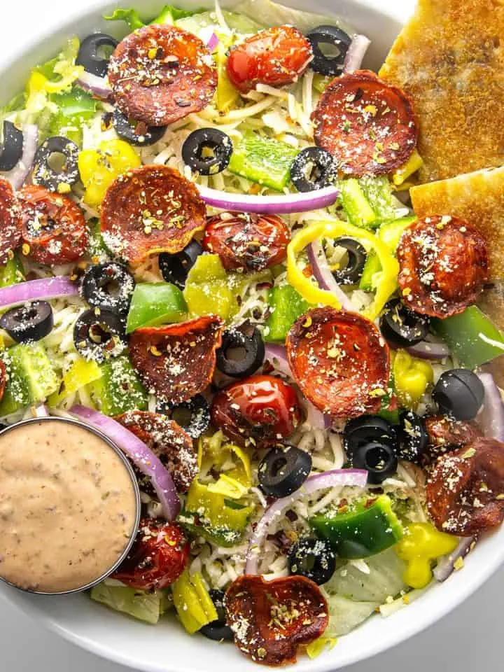 Salad with pepperoni, black olives, cherry tomatoes, bell peppers, red onion, shredded cheese, banana peppers, dressing, and pita wedges in a white bowl.
