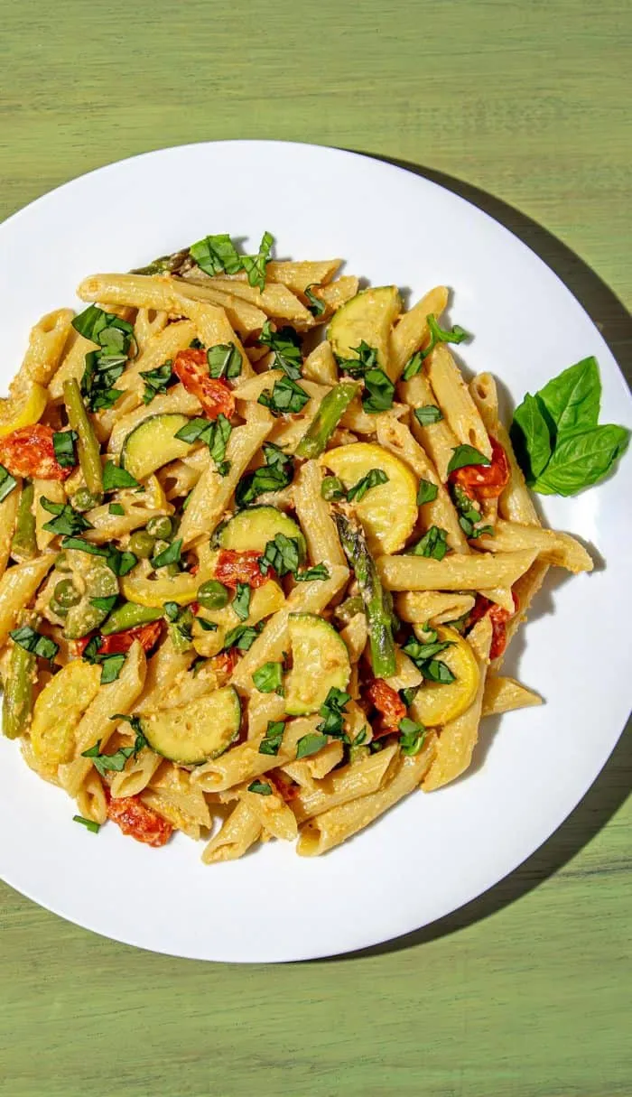 Penne pasta with asparagus, zucchini, cherry tomatoes, peas, and fresh basil on a white plate with a green wooden background.
