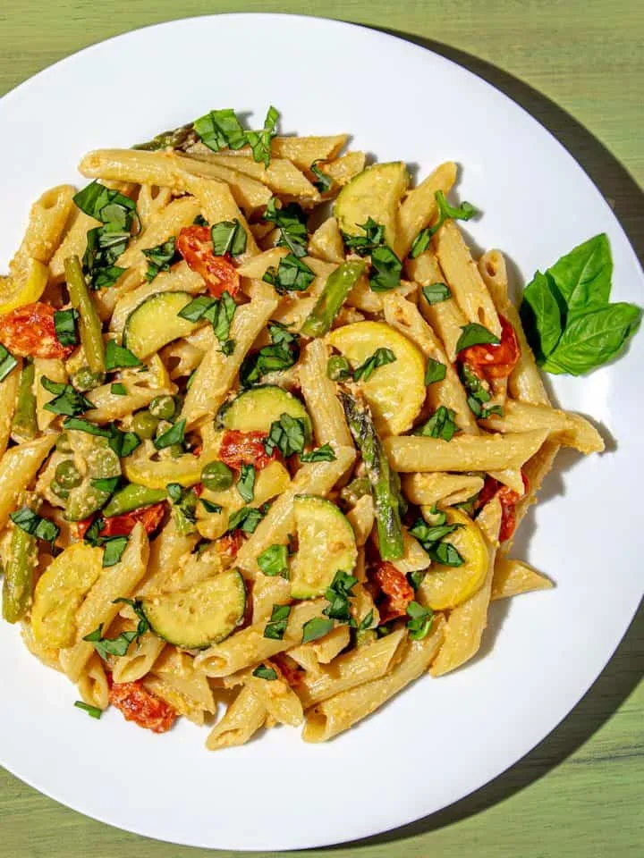 Penne pasta with asparagus, zucchini, cherry tomatoes, peas, and fresh basil on a white plate with a green wooden background.