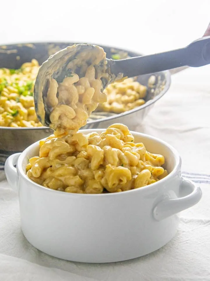 Creamy macaroni and cheese being served from a pan into a white ceramic bowl on a white cloth.