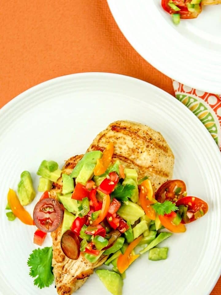 Grilled chicken breast topped with diced avocado, cherry tomatoes, bell peppers, and cilantro on a white plate with an orange background.