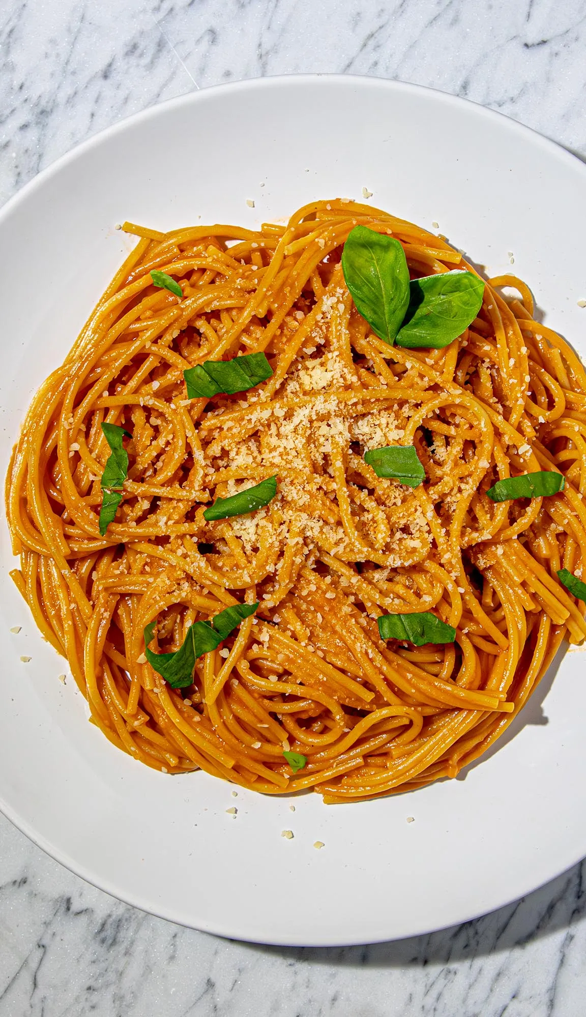 Spaghetti with tomato sauce, sprinkled with grated cheese and fresh basil leaves on a white plate over marble surface.