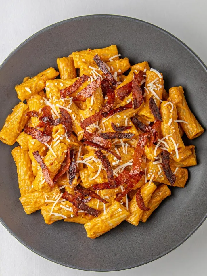 Rigatoni pasta in creamy orange sauce topped with crispy bacon strips and grated cheese on a dark gray plate.
