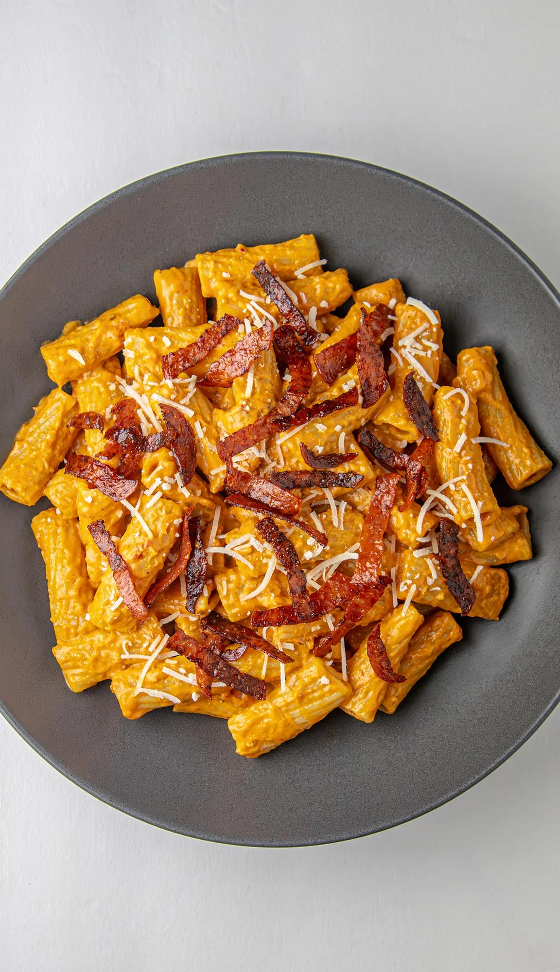 Rigatoni pasta in creamy orange sauce topped with crispy bacon strips and grated cheese on a dark gray plate.