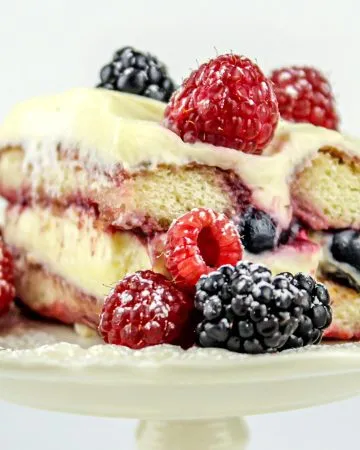 Slice of layered berry cake with cream cheese frosting, topped and surrounded by fresh raspberries and blackberries on a decorative white plate.
