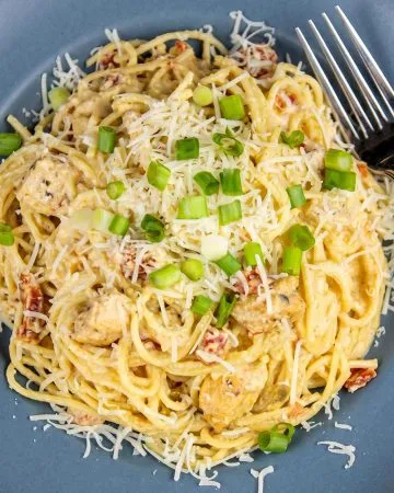 Spaghetti pasta with creamy sauce, chunks of chicken, tomatoes, shredded cheese, and chopped green onions on a blue plate with a fork.