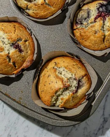 Four freshly baked cherry chia muffins in a metal baking tray on a marble countertop.