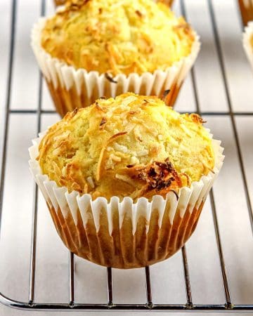 Four golden-brown coconut muffins in white paper liners cooling on a metal rack against a white background.