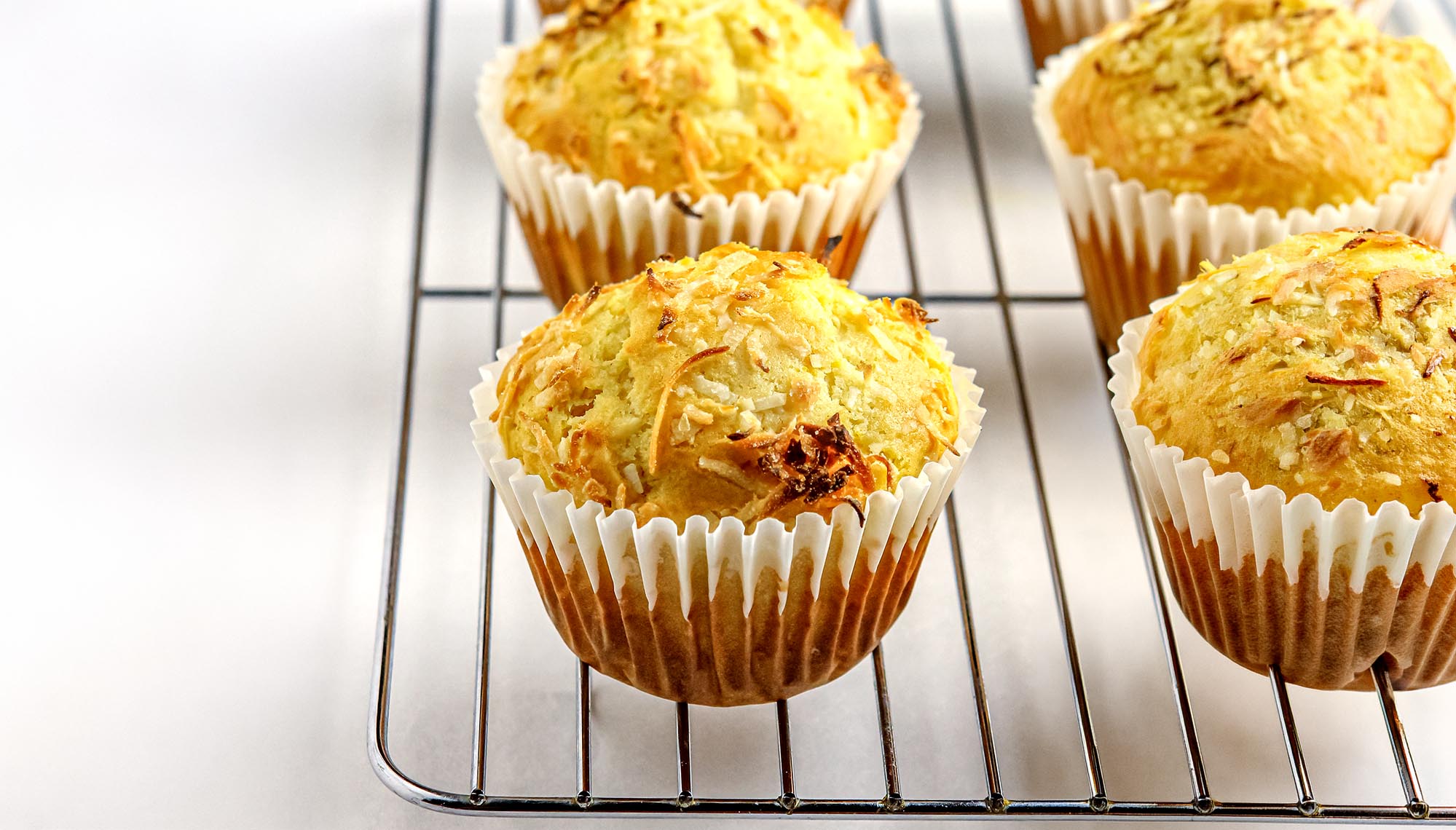 Four golden-brown coconut muffins in white paper liners cooling on a metal rack against a white background.