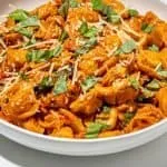 Tortellini pasta in creamy tomato sauce, garnished with shredded cheese and chopped fresh basil in a white bowl on white surface.