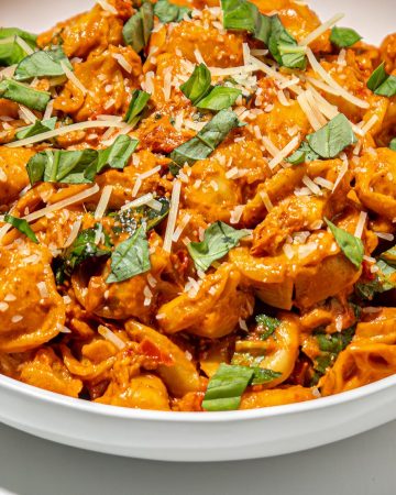 Tortellini pasta in creamy tomato sauce, garnished with shredded cheese and chopped fresh basil in a white bowl on white surface.