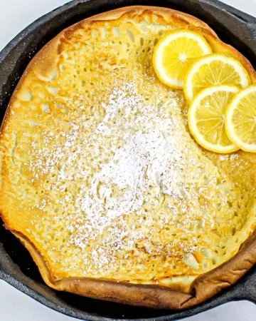 Dutch baby pancake in a cast iron skillet topped with powdered sugar and four lemon slices on a white background.