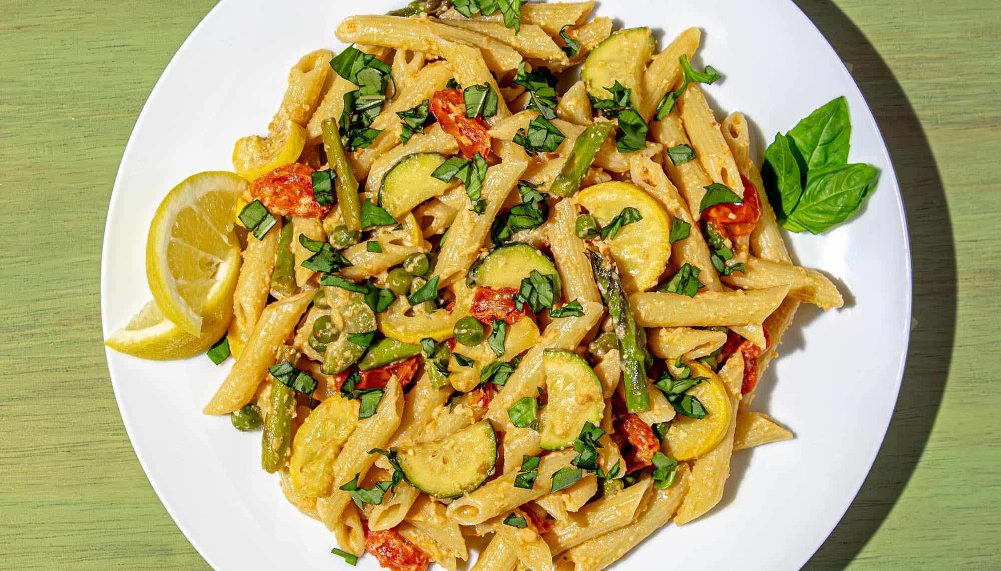 Penne pasta with zucchini, yellow squash, cherry tomatoes, asparagus, peas, fresh basil, and lemon wedges on a white plate.