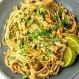 Noodles with chopped peanuts, sliced basil, and two lime wedges on a gray plate.