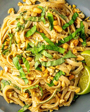 Noodles with chopped peanuts, sliced basil, and two lime wedges on a gray plate.