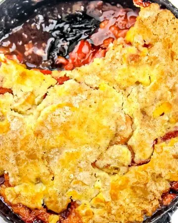 Baked fruit cobbler in a black skillet with a golden-brown crust and bubbling red and dark fruit filling.