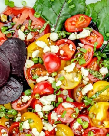 Mixed salad with yellow and red cherry tomatoes, beet slices, leafy greens, chopped green onions, feta cheese, and balsamic glaze on a white plate.
