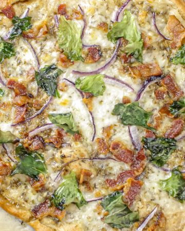 Thin-crust pizza topped with melted cheese, red onion slices, bacon pieces, and spinach leaves on parchment paper.