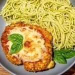 Breaded chicken parmesan topped with melted cheese and basil leaves, served with spaghetti tossed in pesto sauce on a gray plate.