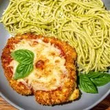 Breaded chicken parmesan topped with melted cheese and basil leaves, served with spaghetti tossed in pesto sauce on a gray plate.