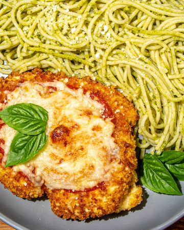 Breaded chicken parmesan topped with melted cheese and basil leaves, served with spaghetti tossed in pesto sauce on a gray plate.