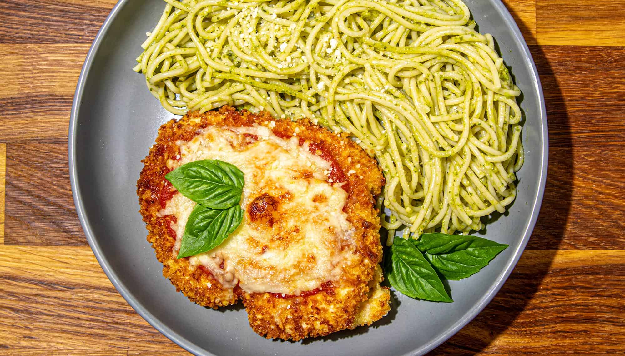 Breaded chicken parmesan topped with melted cheese and basil leaves, served with spaghetti tossed in pesto sauce on a gray plate.
