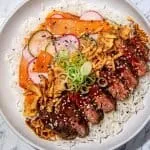 Sliced steak with red thai curry sauce, fried onions, cucumber and radish slices, carrot ribbons, green onions, and sesame seeds on white rice.
