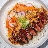 Sliced steak with red thai curry sauce, fried onions, cucumber and radish slices, carrot ribbons, green onions, and sesame seeds on white rice.