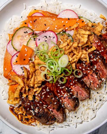 Sliced steak with red thai curry sauce, fried onions, cucumber and radish slices, carrot ribbons, green onions, and sesame seeds on white rice.