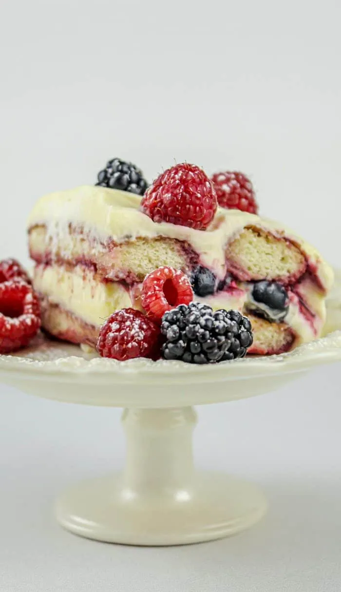 Slice of layered berry tiramisu with raspberries and blackberries on a white pedestal cake stand.