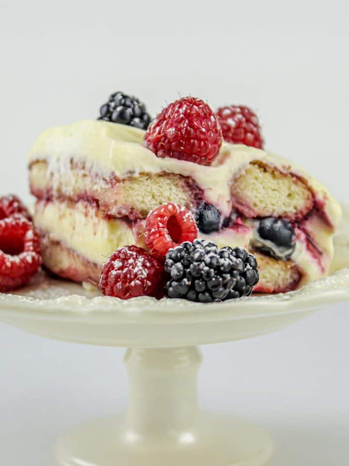 Slice of layered berry tiramisu with raspberries and blackberries on a white pedestal cake stand.
