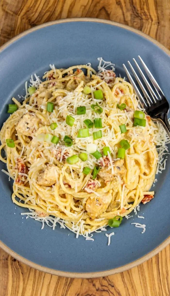 Spaghetti with creamy sauce, chicken chunks, tomatoes, shredded cheese, and chopped green onions on a blue plate with a fork.