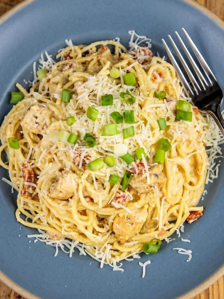 Spaghetti with creamy sauce, chicken chunks, tomatoes, shredded cheese, and chopped green onions on a blue plate with a fork.